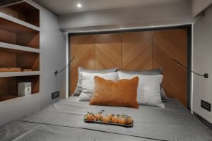 A modern bedroom interior on a Sunreef Yachts vessel features a cozy bed with gray and orange bedding. There are wooden panels on the wall and a shelf in the corner. A tray of tomatoes is placed on the bed.