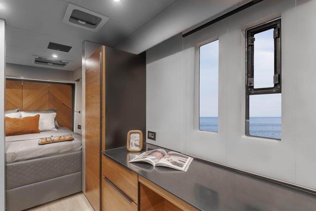 The interior of a Sunreef Yachts vessel, featuring a serene bedroom with a cozy bed and a modern cabinet. An open magazine lies on the cabinet next to a small frame. A window reveals a beautiful sea view.