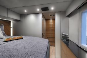 A luxurious cabin interior on a Sunreef Yachts vessel. The room features a large bed adorned with a gray textured cover and a tray of fresh oranges. Warm wooden accents are seen on the walls, and there's a glimpse of the sea through the window.