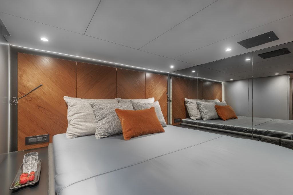 Interior of a Sunreef Yachts cabin featuring a modern and elegant design with a large bed, orange and grey pillows, wood paneling, and a mirrored wall.