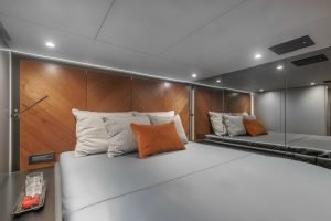 Interior of a Sunreef Yachts cabin featuring a modern and elegant design with a large bed, orange and grey pillows, wood paneling, and a mirrored wall.