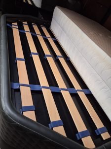 A close-up view of a bed frame with wooden slats held together with blue plastic brackets. Next to it is a mattress. The setting appears indoors, possibly in a storage or assembly area. No relation to Sunreef Yachts is visible.