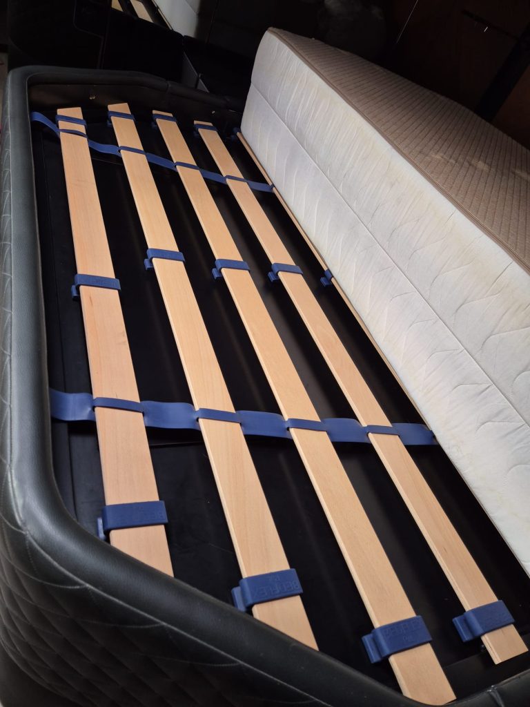 A close-up view of a bed frame with wooden slats held together with blue plastic brackets. Next to it is a mattress. The setting appears indoors, possibly in a storage or assembly area. No relation to Sunreef Yachts is visible.