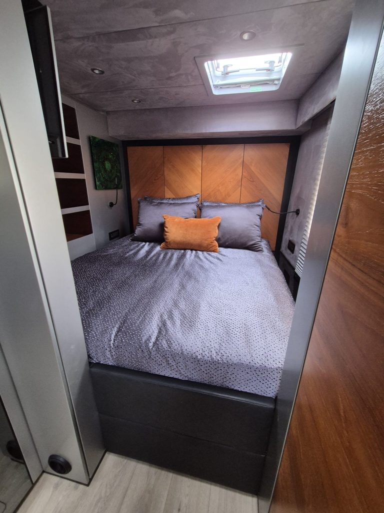 A luxurious cabin interior on a Sunreef Yachts vessel, featuring a plush bed with gray bedding and a brown accent pillow, surrounded by rich wood paneling and a small skylight overhead.