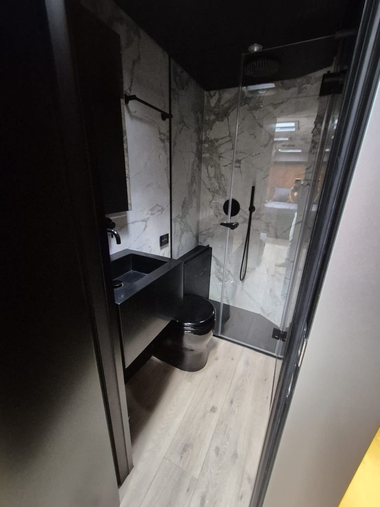 This image shows a modern bathroom in a Sunreef Yachts vessel, featuring a sleek, marble-accented shower, black fixtures, and a wooden floor.