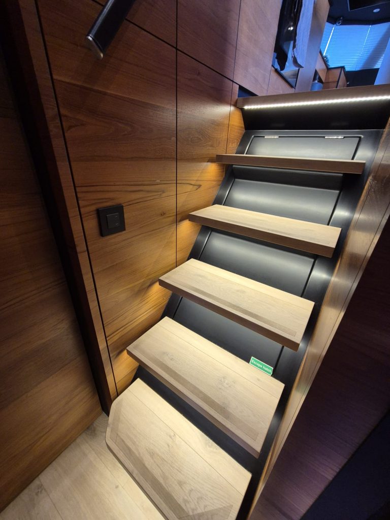 The image shows a modern wooden staircase inside a Sunreef Yachts interior, featuring sleek wooden steps with built-in lighting along the side.