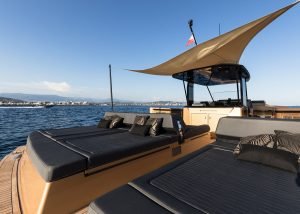 The image shows the deck of a luxury yacht by Sunreef Yachts, featuring black cushioned lounge areas under a sunshade, with a view of the ocean and the coastline in the background.