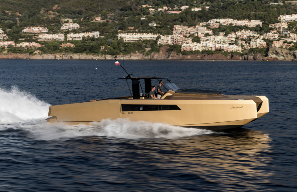 A sleek, golden-colored Sunreef Yachts motor yacht cruising quickly across the water, with scenic hills and buildings in the background.
