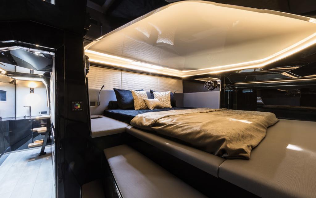 The image shows a luxurious bedroom inside a Sunreef Yachts vessel. The room features a large bed with modern linens and pillows, surrounded by sleek, dark-colored walls and ambient lighting.