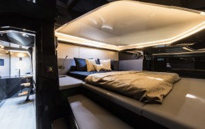 The image shows a luxurious bedroom inside a Sunreef Yachts vessel. The room features a large bed with modern linens and pillows, surrounded by sleek, dark-colored walls and ambient lighting.