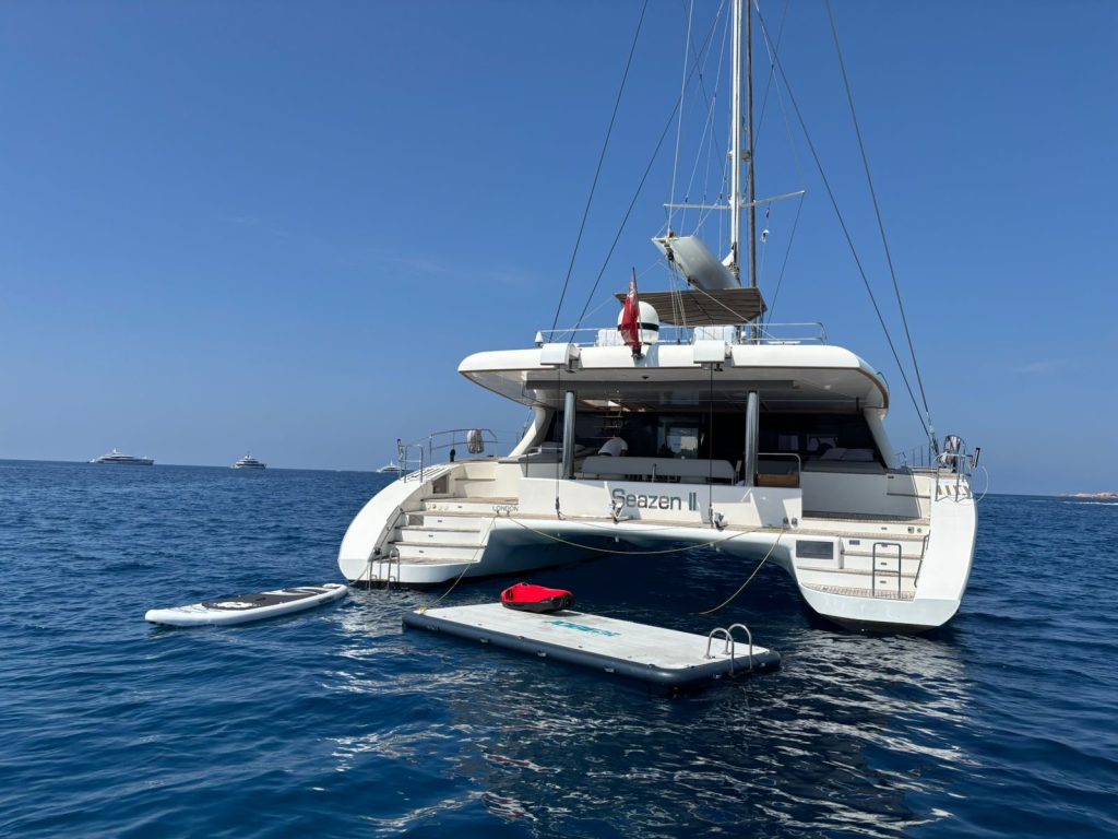 A Sunreef Yachts catamaran named Seazen II is anchored in calm blue water. A person stands on the deck, and a paddleboard and floating platform are nearby.