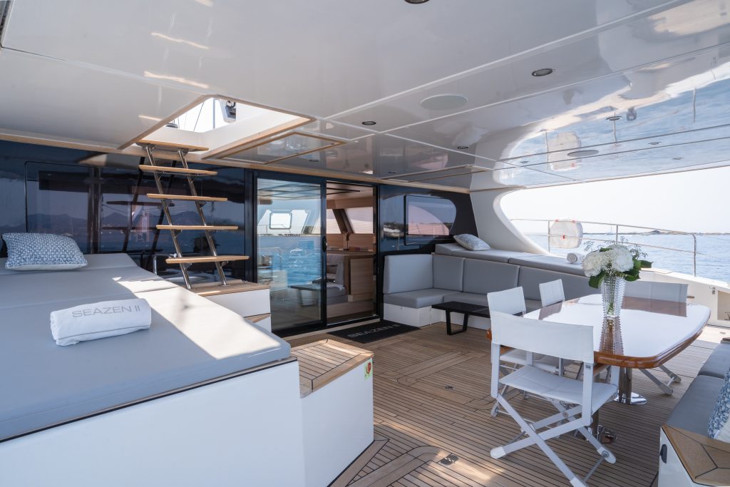 The Sunreef Yachts deck features a luxurious seating area with plush white cushions and a table set with flowers. A staircase leads to an upper level, and large windows offer a view of the ocean.