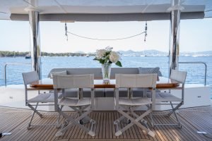 A luxurious dining area on a Sunreef Yachts catamaran, featuring a wooden table with six white chairs. A vase with white flowers sits in the center. Behind, calm waters and a distant hilly coastline are visible.