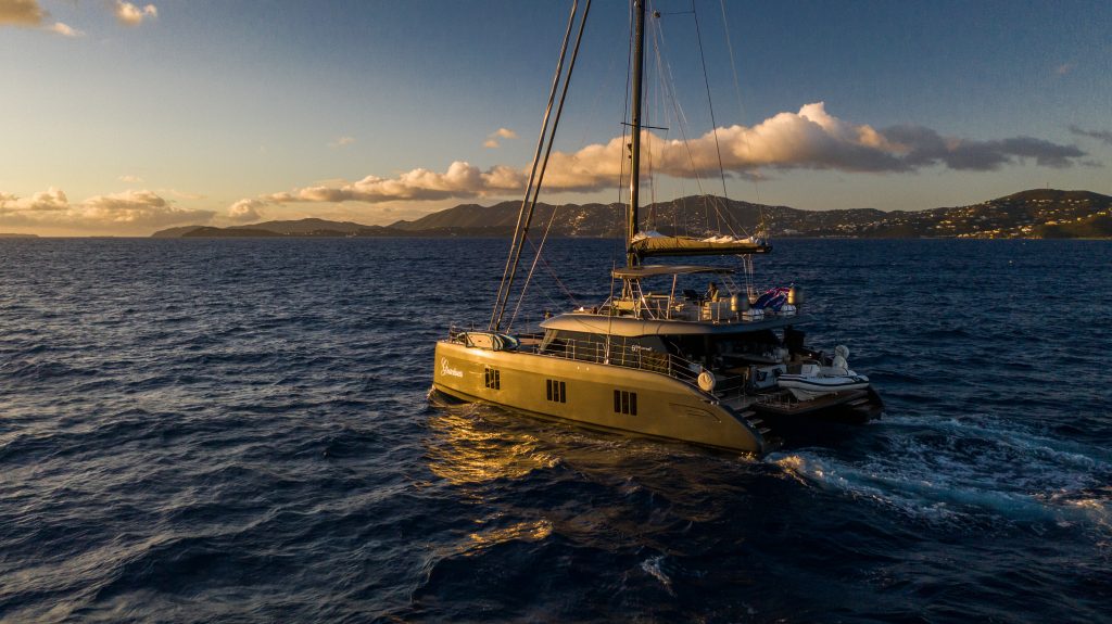 A luxurious Sunreef Yachts catamaran sails in the open sea during sunset. The water glistens under the golden light, and distant islands can be seen on the horizon.