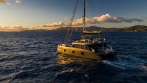 A luxurious Sunreef Yachts catamaran sails in the open sea during sunset. The water glistens under the golden light, and distant islands can be seen on the horizon.