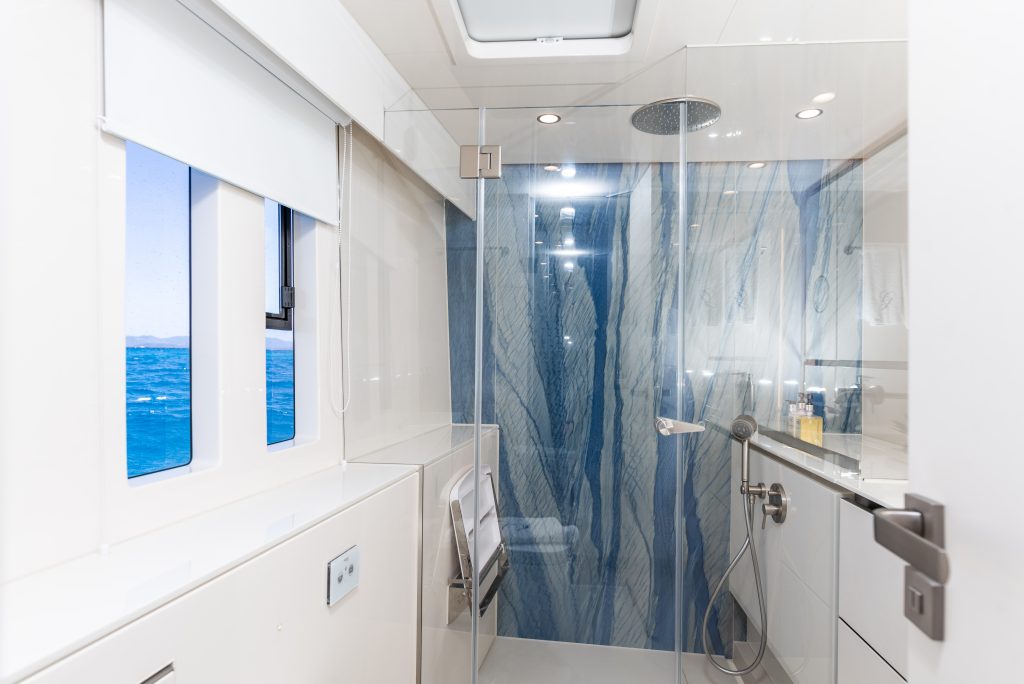 A modern bathroom interior on a Sunreef Yachts vessel features a sleek, glass-enclosed shower with blue-veined marble walls. Large windows provide a view of the ocean, enhancing the luxurious atmosphere.