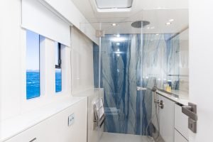 A modern bathroom interior on a Sunreef Yachts vessel features a sleek, glass-enclosed shower with blue-veined marble walls. Large windows provide a view of the ocean, enhancing the luxurious atmosphere.