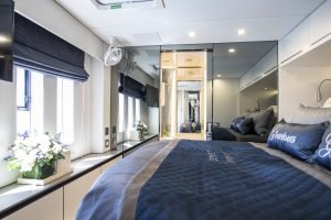 The interior of a luxurious Sunreef Yachts cabin featuring a comfortable bed with elegant bedding, a large mirror, windows with blinds letting in natural light, and decorative flowers on a shelf.