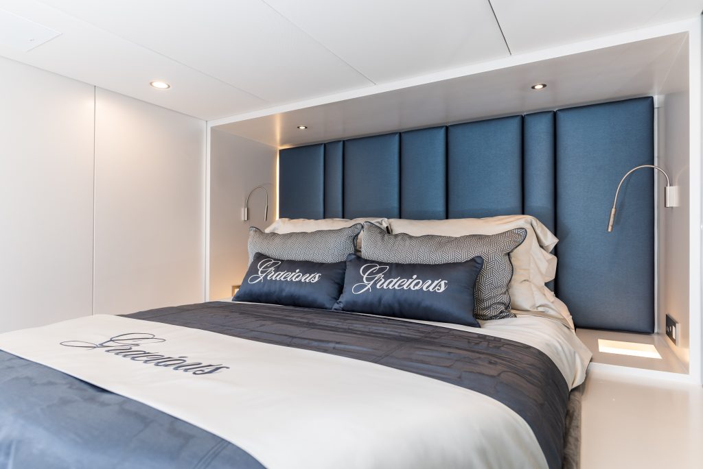 A luxurious bedroom aboard a Sunreef Yachts vessel, featuring an elegant bed with plush blue headboard and bedding, and pillows embroidered with the word 'Gracious'.
