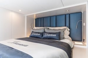 A luxurious bedroom aboard a Sunreef Yachts vessel, featuring an elegant bed with plush blue headboard and bedding, and pillows embroidered with the word 'Gracious'.