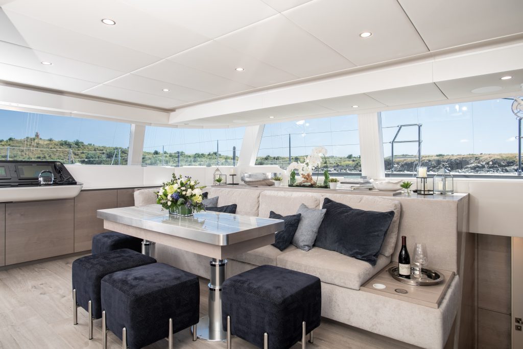 Interior view of a Sunreef Yachts catamaran, showcasing a luxurious and modern living space with large windows, a stylish dining area, plush seating with cushions, and decorative plants and flowers.