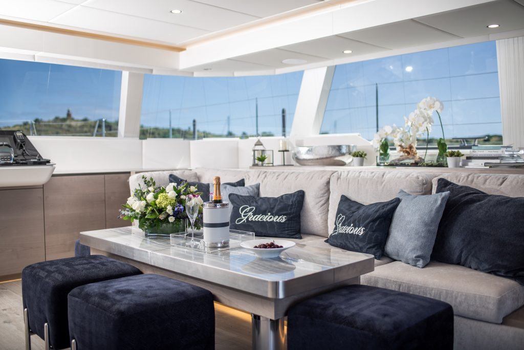 The interior of a luxury yacht by Sunreef Yachts, featuring a plush seating area with 'Gracious' embroidered pillows, a sleek table with a champagne bottle, and large windows offering a scenic view.