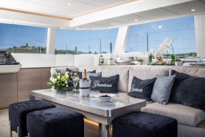 The interior of a luxury yacht by Sunreef Yachts, featuring a plush seating area with 'Gracious' embroidered pillows, a sleek table with a champagne bottle, and large windows offering a scenic view.