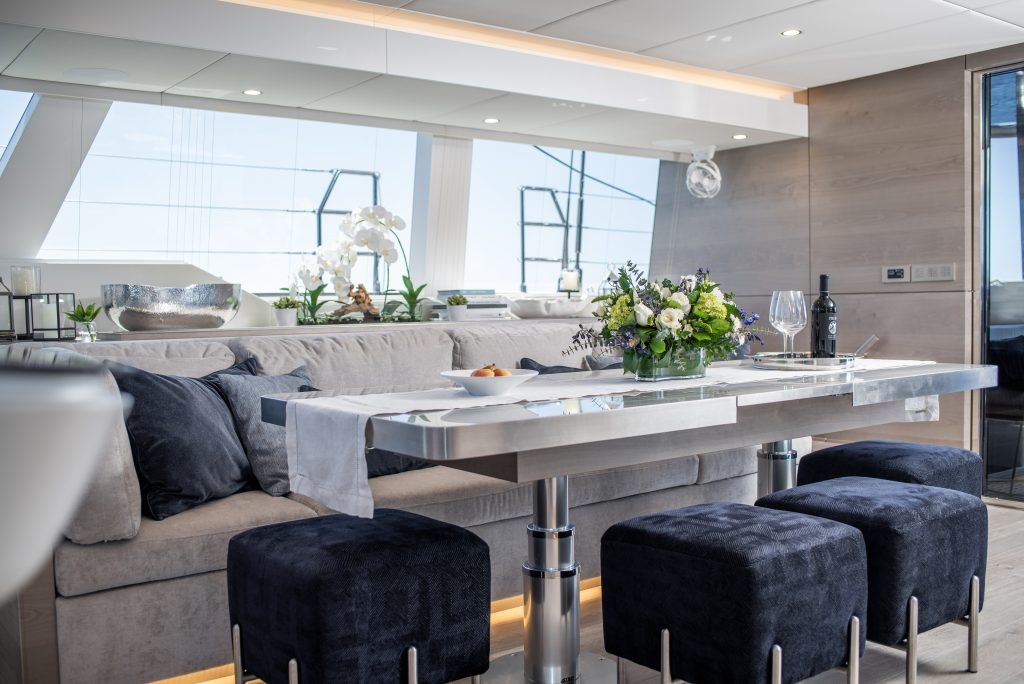 Interior of a luxury yacht by Sunreef Yachts, featuring a sleek dining area with a modern metal table, comfortable blue cushioned stools, and a gray sofa. The table is elegantly set with flowers and glassware.