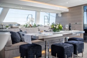 Interior of a luxury yacht by Sunreef Yachts, featuring a sleek dining area with a modern metal table, comfortable blue cushioned stools, and a gray sofa. The table is elegantly set with flowers and glassware.