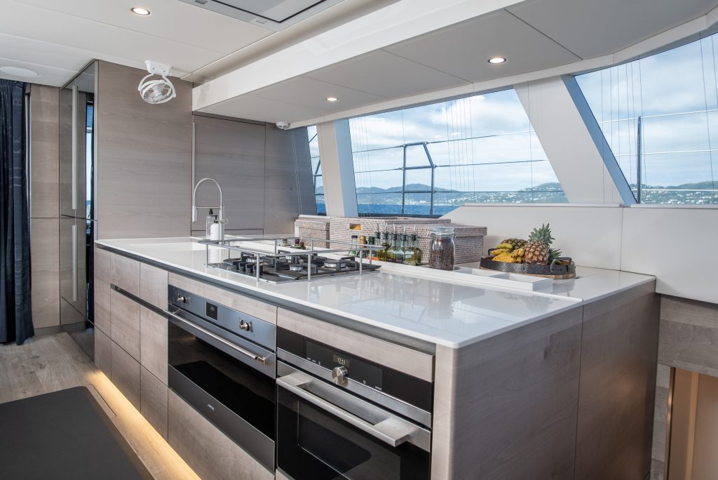 The interior of a Sunreef Yachts vessel features a modern kitchen with a stove, oven, and sleek countertops. The large windows provide a view of the sea.