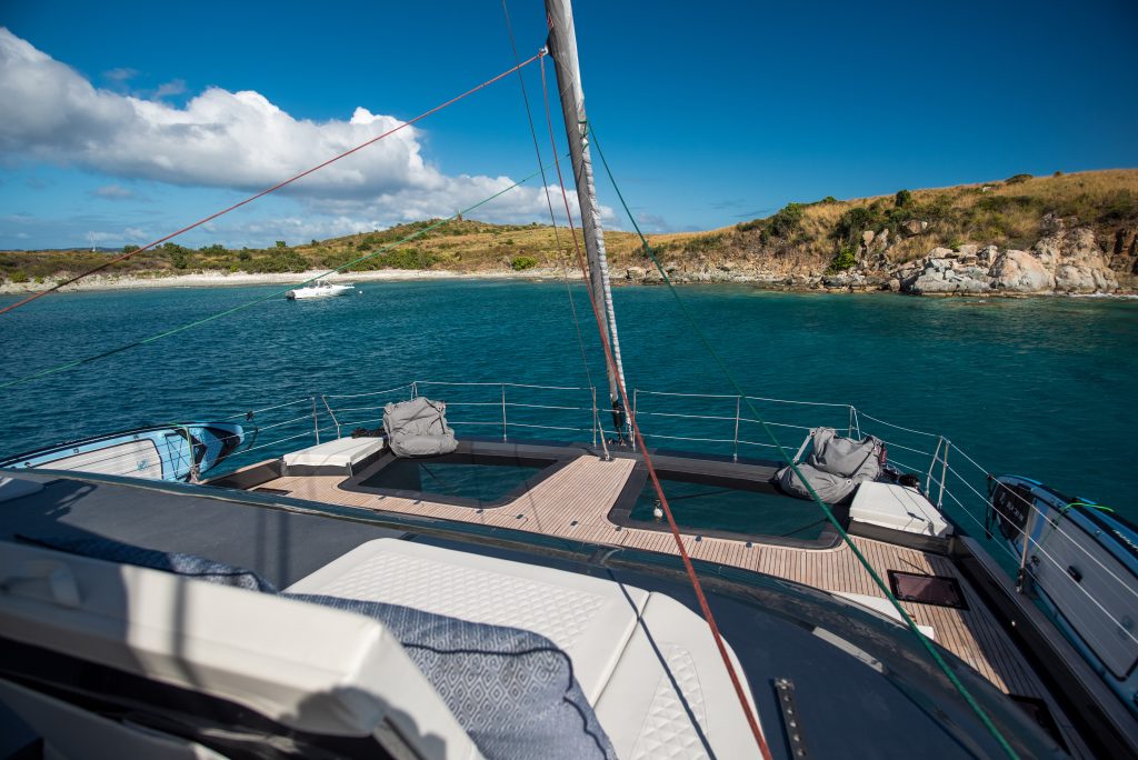 View of Sunreef Yachts sailing catamaran's deck with lounging areas, surrounded by clear blue water and rocky coastline under a bright sky.