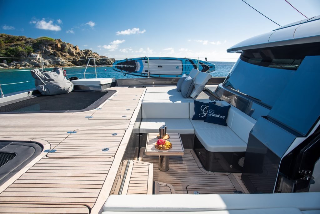 The deck of a Sunreef Yachts catamaran is shown with luxurious seating and a table holding a fruit platter. Paddleboards are mounted above, with a scenic ocean view in the background.