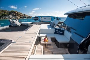 The deck of a Sunreef Yachts catamaran is shown with luxurious seating and a table holding a fruit platter. Paddleboards are mounted above, with a scenic ocean view in the background.