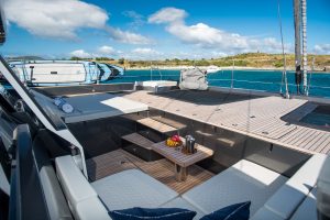 The image shows the deck of a luxurious Sunreef yacht. It features comfortable seating areas with cushions, a small table set with fruit and drinks, and a clear blue sky with some clouds. The yacht is anchored near a scenic coastline.