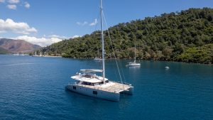 A Sunreef Yachts catamaran sailing on a calm blue sea with a forested hillside in the background.