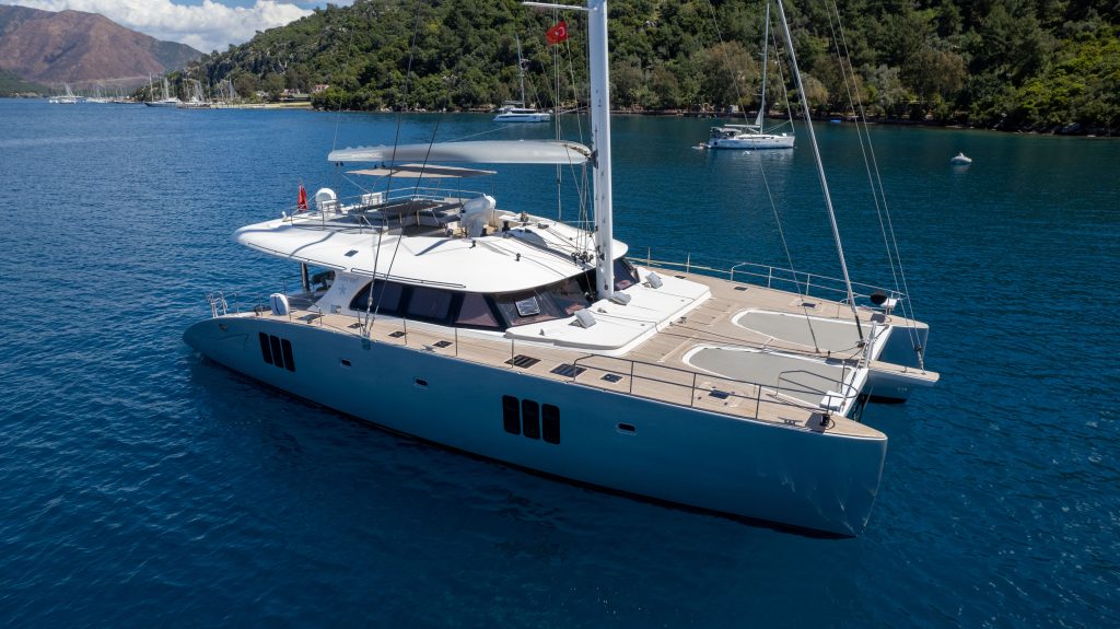 A Sunreef Yachts catamaran is anchored on a calm blue sea, surrounded by lush green hills and other boats in the distance.