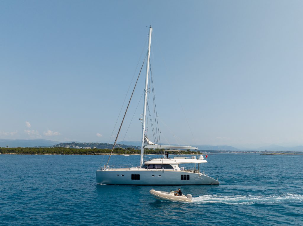 A Sunreef Yachts catamaran sails on a blue ocean with a small inflatable boat nearby.