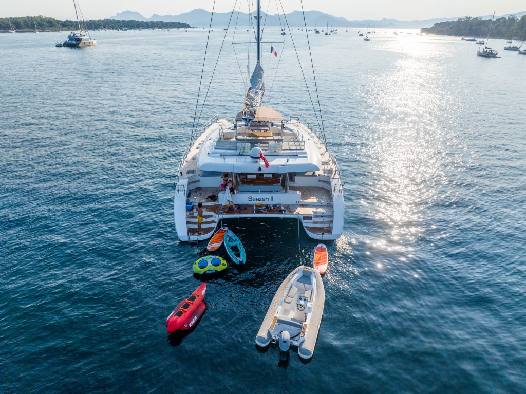 A catamaran made by Sunreef Yachts is anchored in the water, surrounded by small boats and inflatable watercraft, with a sunny landscape in the background.