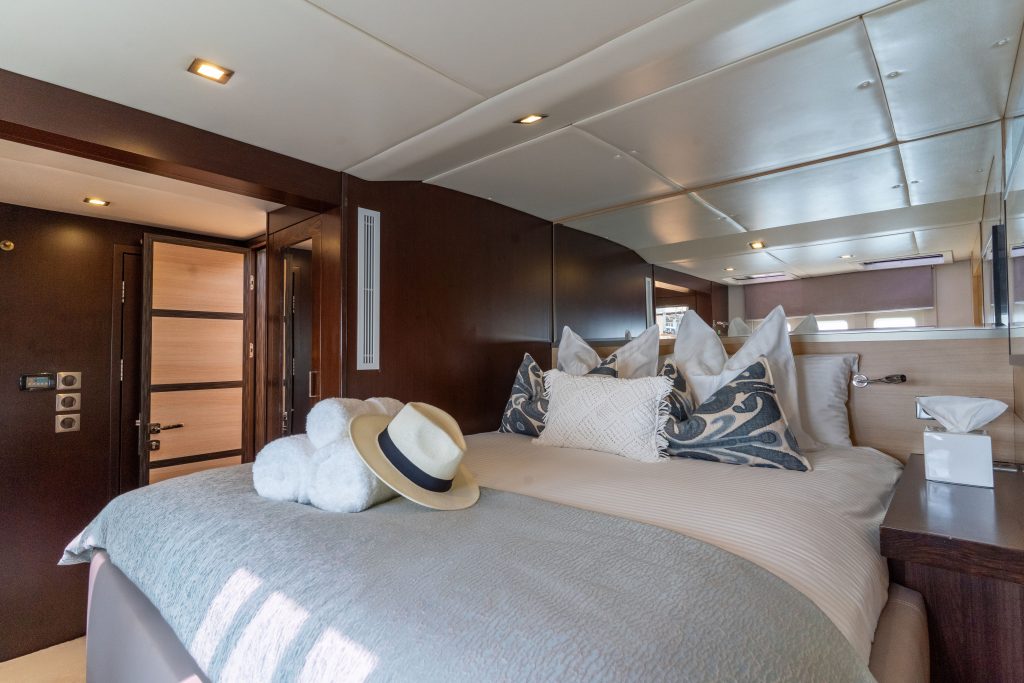 Interior view of a luxurious bedroom on a Sunreef Yachts boat, featuring a large, comfortable bed with decorative pillows, a straw hat placed on the bed, and warm ambient lighting.