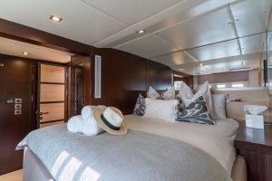 Interior view of a luxurious bedroom on a Sunreef Yachts boat, featuring a large, comfortable bed with decorative pillows, a straw hat placed on the bed, and warm ambient lighting.