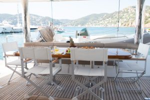 The deck of a Sunreef Yachts catamaran features a dining setup with a wooden table surrounded by white folding chairs. The table is adorned with fruits and glasses, and offers a view of the marina with other boats and a scenic hilly landscape in the background.