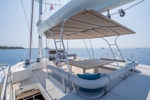 The image shows the deck of a luxurious Sunreef Yachts sailing boat. It features a seating area with a table beneath a canopy, with a serene ocean view in the background.
