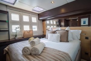Interior of a luxury Sunreef Yachts cabin featuring a neatly made bed with white and beige linens, three rolled towels, and decorative pillows. A window allows natural light in, with a plant and a shopping bag also in view.