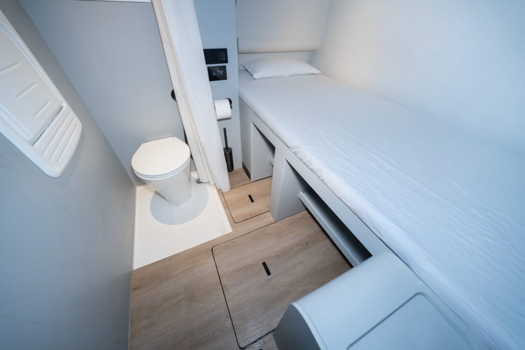 The image shows a compact cabin interior featuring simple furnishings, including a bed with white linens and a toilet, onboard a Sunreef Yachts vessel.