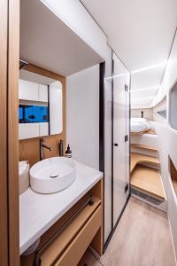 Interior of a Sunreef Yachts cabin featuring a modern bathroom with a round sink and a wooden cabinet. A sleeping area with a bed is visible in the background.