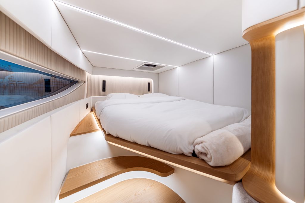 The image shows the interior of a Sunreef Yachts cabin. It features an elegantly designed bed with white bedding and a wooden frame. The cabin is illuminated with soft lighting and has a modern, minimalist design with a window on the left side.