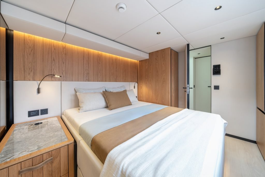 Interior cabin of a Sunreef Yachts catamaran, featuring a double bed with beige and brown bedding, wooden paneling, and modern lighting.