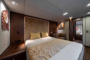 A luxurious Sunreef Yachts cabin interior showcasing a cozy double bed with patterned bedding, wooden accents, and dim lighting. Decor includes artistic wall pieces, and a small corner seating area is visible with cushions.