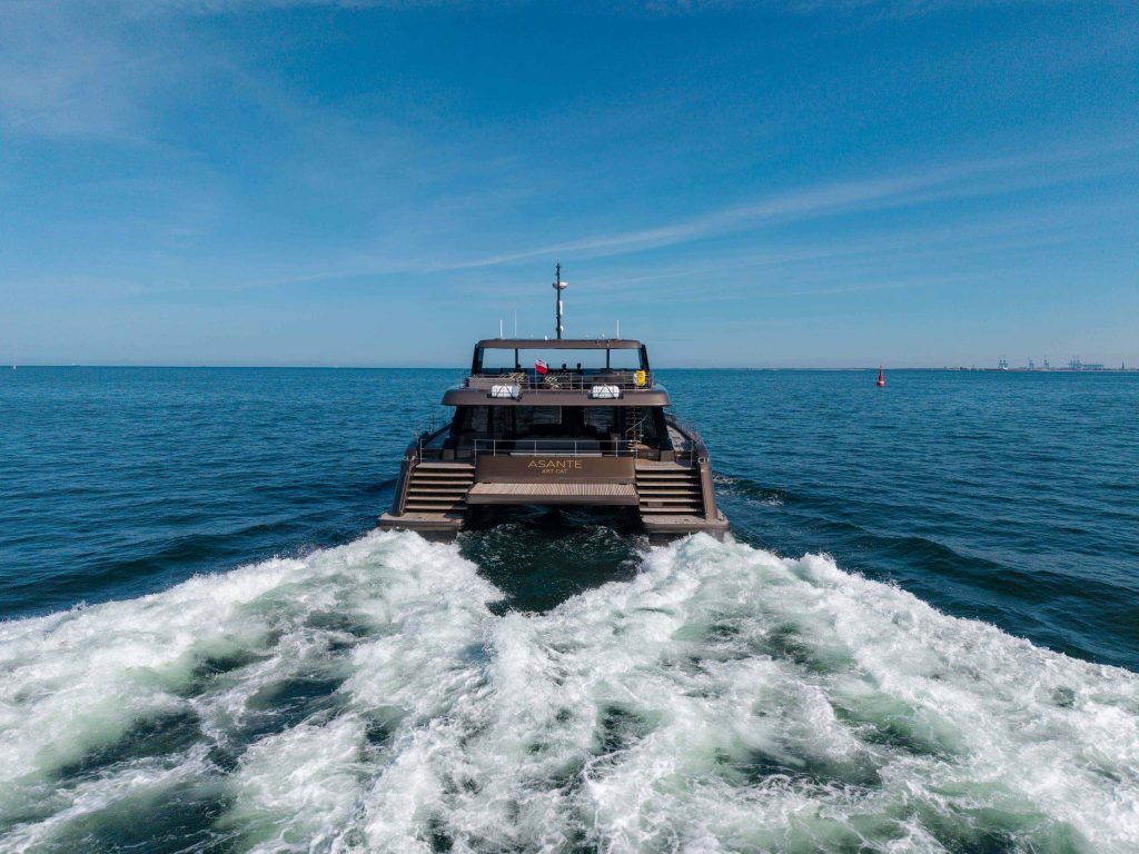 A Sunreef Yachts catamaran named Aganippe cruises on the open sea, leaving a foamy wake behind under a clear blue sky.
