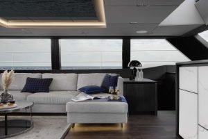 Interior of a Sunreef Yachts vessel showcasing a modern, elegant lounge area. The space features a comfortable gray sofa with plush cushions, a round coffee table adorned with decorative elements, and subtle lighting. Large windows offer a clear view of the sea, enhancing the luxurious ambiance.